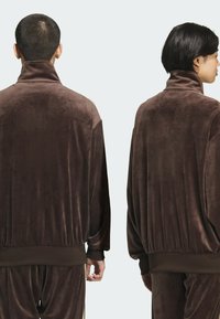 Brown velour track jacket featuring a high collar, dropped shoulders, and elastic cuffs. Smooth texture with a relaxed fit and minimal detail.