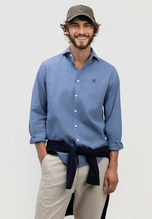 Hombre sonriente que lleva una gorra verde, camisa azul abotonada con las mangas remangadas, pantalones beige y un suéter oscuro atado alrededor de la cintura.