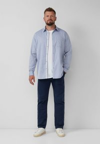Lichtblauw button-up shirt met een borstzak, gedragen over een wit T-shirt; gecombineerd met donkerblauwe broek en witte sneakers.