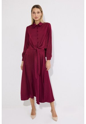 Žena u dugoj haljini na kopčanje, tamnocrvena midi haljina s vezanim strukom, stoji ispred jednobojne bijele pozadine.