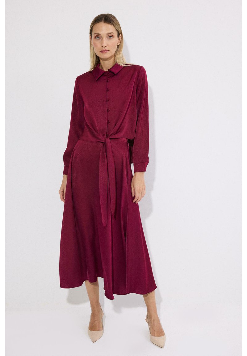 Γυναίκα που φορά ένα μακρυμάνικο, κουμπωτό, βαθύ κόκκινο μίντι φόρεμα με δεμένη μέση, στέκεται μπροστά σε ένα απλό λευκό φόντο.
