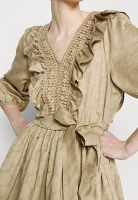 Robe beige en tissu texturé avec un décolleté en V, des volants, des détails en dentelle complexes et une taille ceinturée avec un nœud.