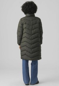 Olijfgroene gewatteerde jas met chevron-quilting, hoge kraag en lange mouwen, gecombineerd met blauwe wijdere jeans en zwarte schoenen.