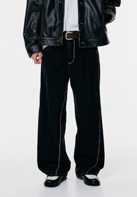 Person iført sort læderjakke, hvid skjorte, løse sorte bukser med synlige syninger, samt sort-hvide sko, der står mod en hvid baggrund.