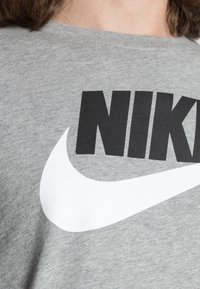Detailní záběr na šedou látku s velkým černým textem "NIKE" a bílým logem Nike swoosh na oděvu.
