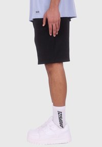 Store bukser med ribbet tekstur, båret med hvide sneakers og hvide sokker med sort tekst. Sidevisning, der viser hånden hvilende på bukserne.