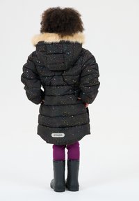 Barn med krøllet hår iført en sort pufferjakke med pelskrave, lilla leggings og sorte gummistøvler, stående med ryggen til.