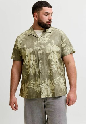 Homme avec une barbe portant une chemise verte à manches courtes avec un léger motif floral et un pantalon gris, regardant vers la gauche.