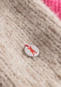 Beige gebreide stof met een zachte textuur heeft een zilveren ronde label met een rode honden silhoette en een rode draad.