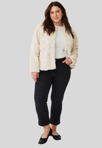 Vrouw in een crèmekleurige fuzzy jas over een witte top, zwarte cropped jeans en zwarte flats, staand met één hand in de zak tegen een grijze achtergrond.