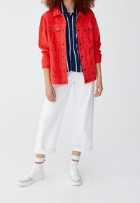 Veste en velours côtelé rouge portée sur une chemise à rayures bleu marine et blanches, associée à un pantalon blanc retroussé et des baskets blanches avec des touches grises.