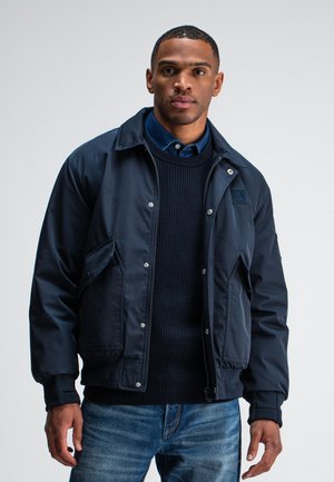 Marineblaue Bomberjacke mit Druckknöpfen, Seitentaschen und gerippten Bündchen, getragen über einem dunklen Strickpullover und einem Jeanshemd.