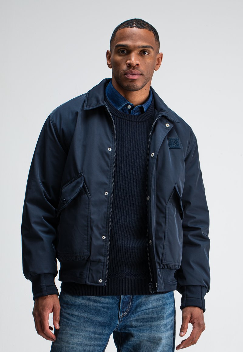 Marineblaue Bomberjacke mit Druckknöpfen, Seitentaschen und gerippten Bündchen, getragen über einem dunklen Strickpullover und einem Jeanshemd.
