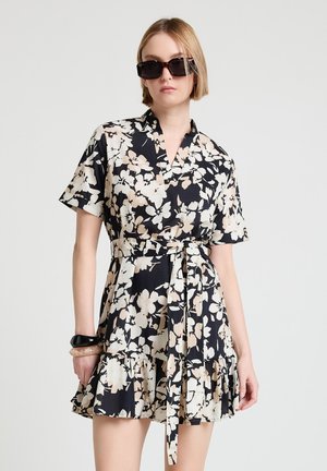 Donna che indossa un vestito corto con maniche a sbuffo a fiori nero e beige con cintura, grandi occhiali da sole neri e due braccialetti al polso sinistro.