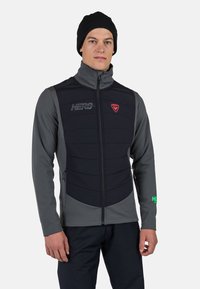 Herrarnas vadderade väst i grått och svart med hög krage, dragkedjestängning, bröstlogo och grön detalj på ärmen. Mjukt, texturerat material.