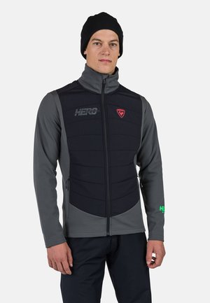 Men's padded vest in gray and black with a high collar, zip closure, chest logo, and green accent on the sleeve. Soft, textured material.