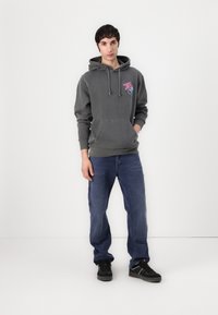 Sweatshirt cinza escuro com bolso canguru e cordão, apresentando um gráfico rosa. Usado com calças de carregador azuis e ténis pretos.