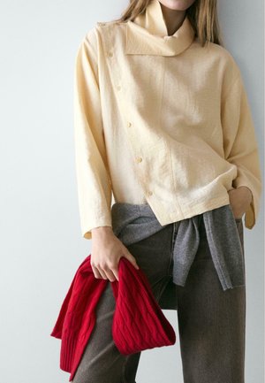 Cream-colored buttoned top with a high collar, paired with gray pants and a red, ribbed knit item held in hand.