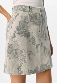 Jupe en denim floral de couleur beige clair avec des motifs floraux gris, dotée de poches latérales et d'une fermeture à boutons. Silhouette classique en A.