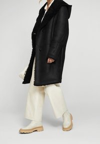 Abrigo de borreguillo negro con capucha, que presenta un gran collar, diseño de doble botonadura y una textura suave. Combinado con pantalones de pierna ancha color crema y botas.