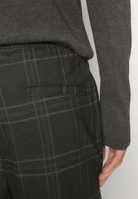 Zwarte geruite broek met grijze lijnen, voorzien van een achterzak en een aansluitende tailleband, gecombineerd met een donkergrijze gebreide trui.