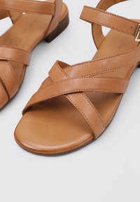 Sandalias de cuero marrón con tiras entrelazadas y un tacón bajo. Textura suave con un detalle de costura mínimo y un diseño de punta redondeada.