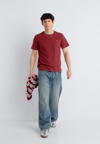 Νεαρός άνδρας που περπατά, φοράει κόκκινη μπλούζα, φαρδιά μπλε τζιν, ασημένια αθλητικά παπούτσια και κρατάει ένα κόκκινο-λευκό ριγέ πουλόβερ.