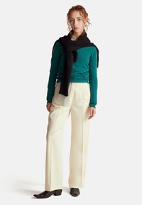 Jersey turquesa, bufanda negra caída sobre los hombros, pantalones blancos de pierna ancha y zapatos negros de punta; texturas suaves y ropa en capas.