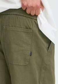 Hand lifting white shirt to reveal elastic waist olive green pants with back pocket and small brand tag.
