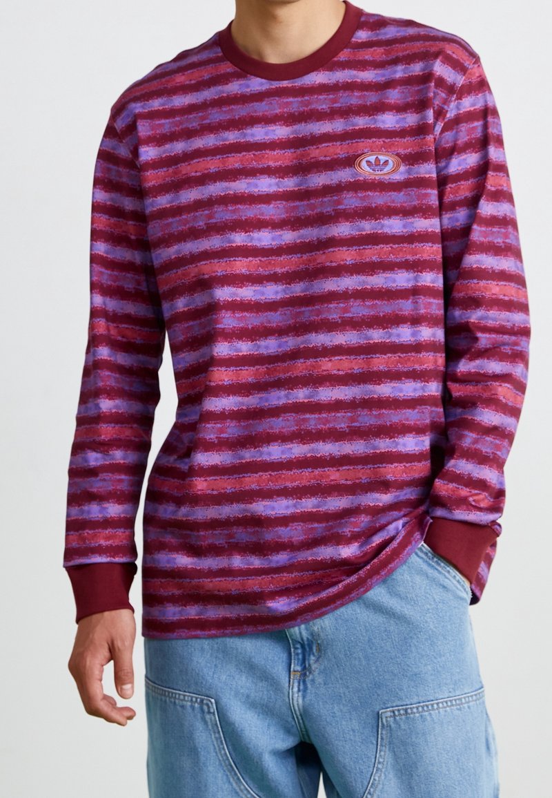 Czerwona bluzka z długim rękawem w poziome fioletowe paski, okrągły dekolt i ściągacze przy rękawach, z małym haftowanym logo na piersi.