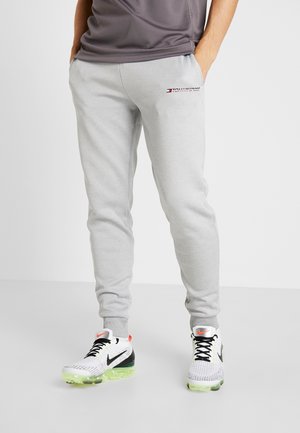 Osoba ubrana w jasnoszare spodnie dresowe z małym logo na udzie i białe buty Nike z neonowo zielonymi podeszwami i czarnymi sznurowadłami.
