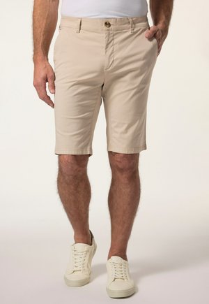 Mann trägt beige knielange Shorts, weiße Turnschuhe und ein weißes Hemd, mit einer Hand in der Tasche, steht vor einem schlichten Hintergrund.