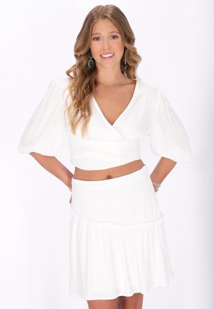 Kvinde med langt bølget hår, smilende, iført hvid crop top med pufærmer og matchende nederdel, stående mod en hvid baggrund.