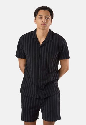 Ung mand stående med hænderne bag ryggen, iført sort kortærmet skjorte med knapper og matchende stribede shorts mod en hvid baggrund.