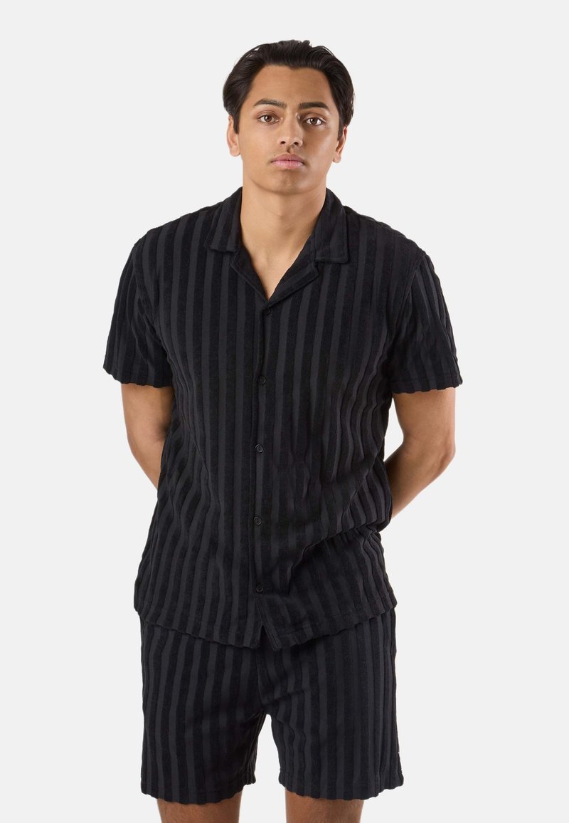 Jeune homme debout, les mains derrière le dos, vêtu d'une chemise noire à manches courtes boutonnée et d'un short assorti rayé sur fond blanc.