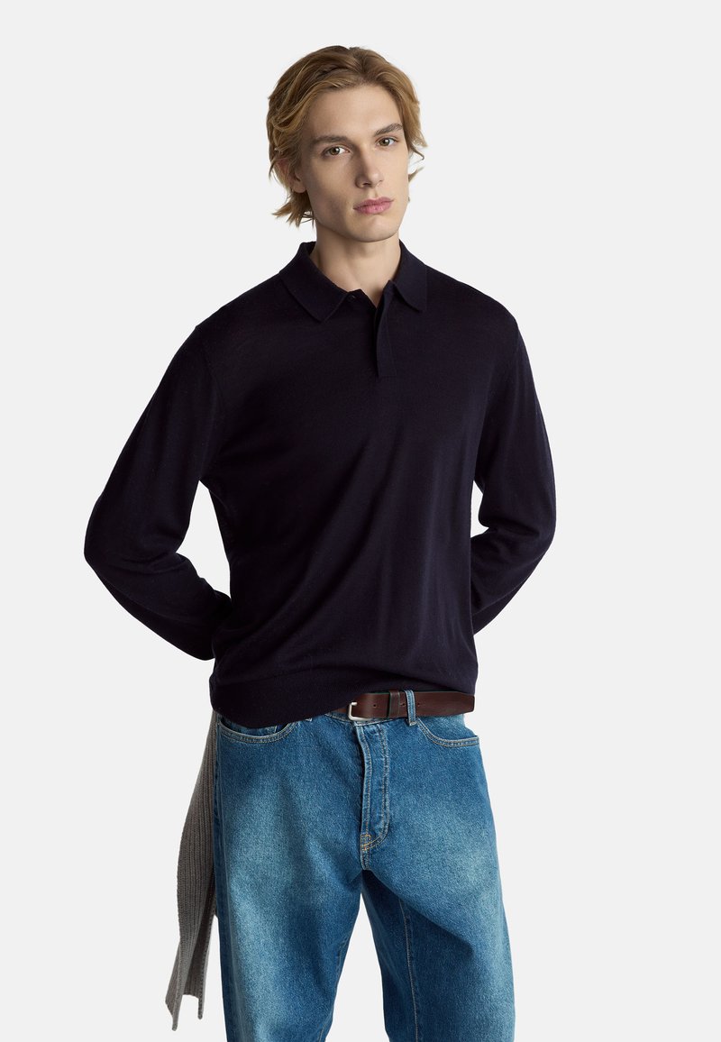 Polo azul marino tejido con mangas largas, textura suave y cuello, combinado con jeans de mezclilla azul claro y un cinturón marrón.