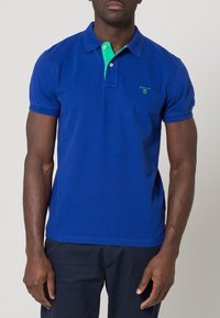 Blå polotröja i bomull med grön krage. Har en liten grön logotyp på bröstet och korta ärmar. Enkelt mönster.