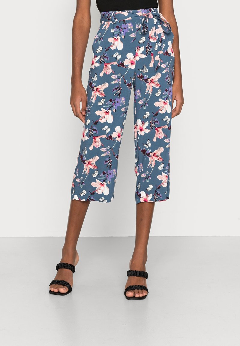 Femme portant un pantalon court bleu à fleurs avec une taille à nouer et des sandales noires à talons hauts, debout devant un fond uni.
