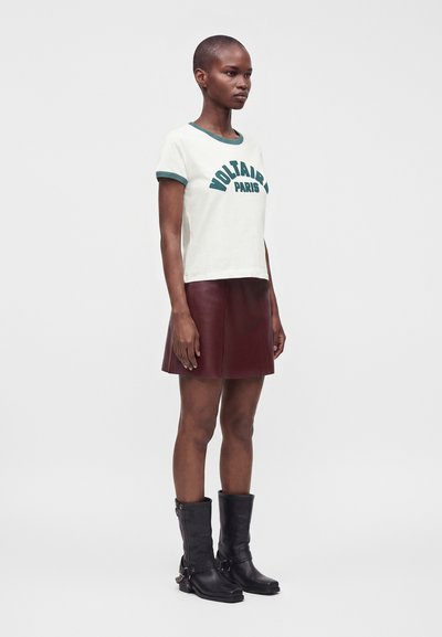 Γυναίκα φοράει λευκό T-shirt με πράσινο περίγραμμα και κείμενο, σκούρα κόκκινη δερμάτινη φούστα και μαύρες μπότες μέχρι τη μέση της γάμπας, στέκεται μπροστά σε απλό φόντο.