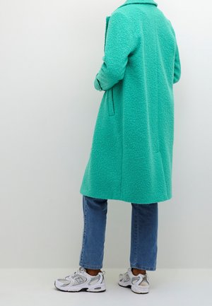 Turkis fleecejakke med krave, lange ærmer og sidelommer; parret med lyseblå jeans og hvide sneakers.