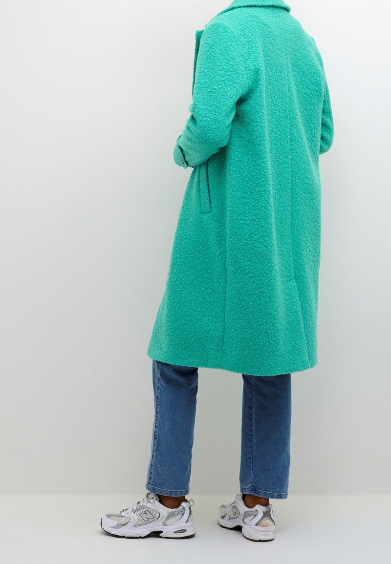 Cappotto in fleece turchese con colletto, maniche lunghe e tasche laterali; abbinato a jeans azzurri e sneakers bianche.