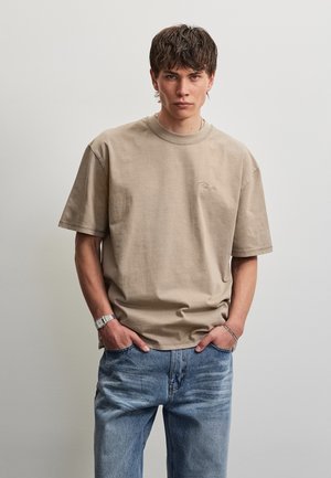 Jeune homme portant un t-shirt beige oversize et un jean bleu, debout les mains dans les poches devant un fond clair uni.