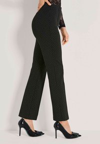 Vrouw in zwarte hoge taille broek met kleine witte stippen en zwarte punkachtige hoge hakken met strikken, staande in zijaanzicht.