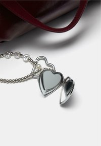 Oglindă compactă în formă de inimă, argintie, cu un lanț decorat cu mărgele și un accent de perlă, așezată lângă o geantă din piele sintetică burgundy.