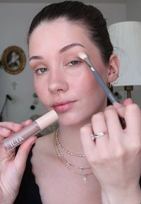 ILIA Make-up-Produkt, das in einer Hand gehalten wird; zylindrischer Behälter mit einem beigen Deckel. Mit einem kleinen Pinsel in der anderen Hand wird Make-up aufgetragen.