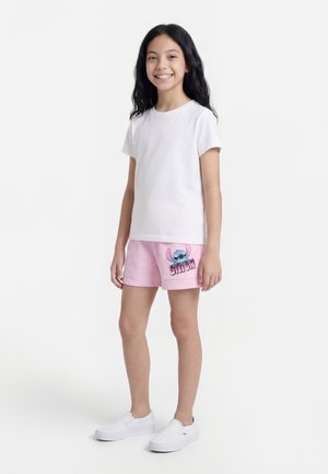 Egy lány áll mosolyogva, fehér pólót, rózsaszín rövidnadrágot, amelyen a Stitch karakter látható, és fehér slip-on cipőt visel, fehér háttér előtt.