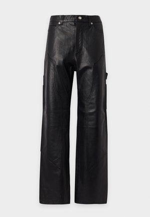 Une femme présente, sur un fond uni, un haut noir sans manches à motif graphique, un pantalon large, une ceinture cloutée, un sac à l’épaule et des bottines.