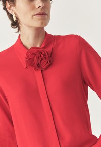 Blusa roja con cuello que presenta una gran rosa de tela. El material parece suave y ligero, complementado por unos pendientes texturizados en dorado.