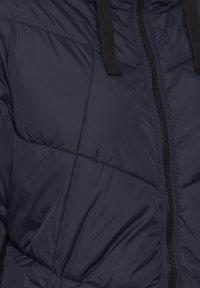 Jachetă pufoasă de culoare bleumarin, cu un design matlasat, fermoar negru și cordon la glugă, realizată dintr-un material neted și lucios.