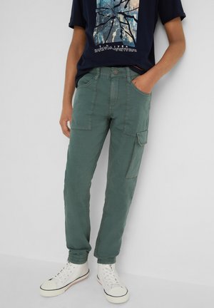 Persona che indossa pantaloni cargo verdi con polsini elastici, una t-shirt grafica blu navy e sneakers bianche alte, in piedi contro un muro bianco.